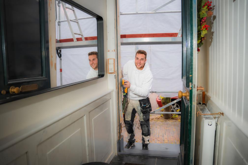 Schilder schuurt een deurpost in een woning en kijkt glimlachend naar de camera